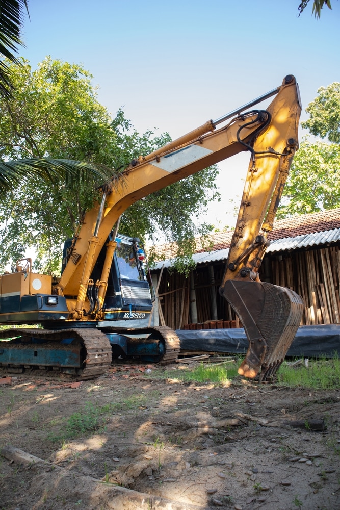 Excavatrice Kobelco jaune et bleue sur chenilles, la flèche levée et le godet au sol, devant un hangar en bois et des arbres verts.
