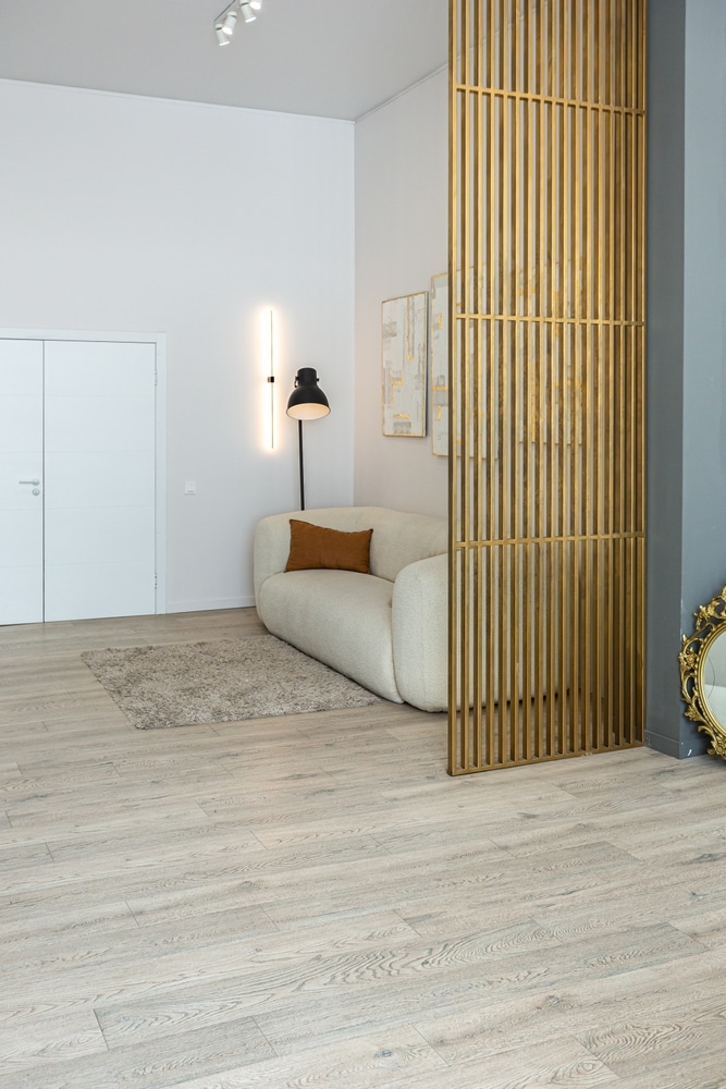 Séparation de salon par claustra à lattes or. Canapé moderne incurvé blanc, sol en bois clair et éclairages design.