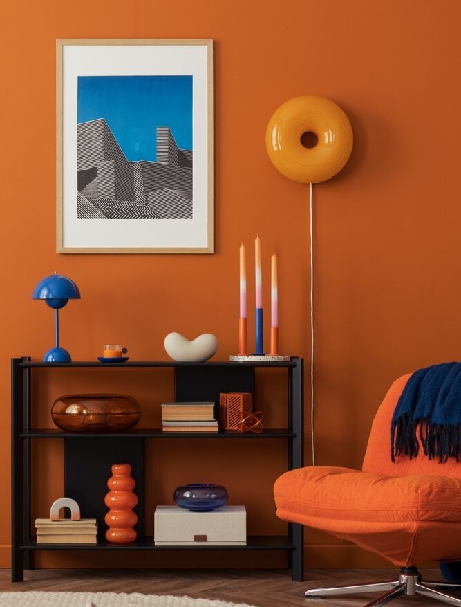 Décoration moderne audacieuse : mur orange, étagère noire, fauteuil orange, lampe bleue et œuvre d'art graphique encadrée.