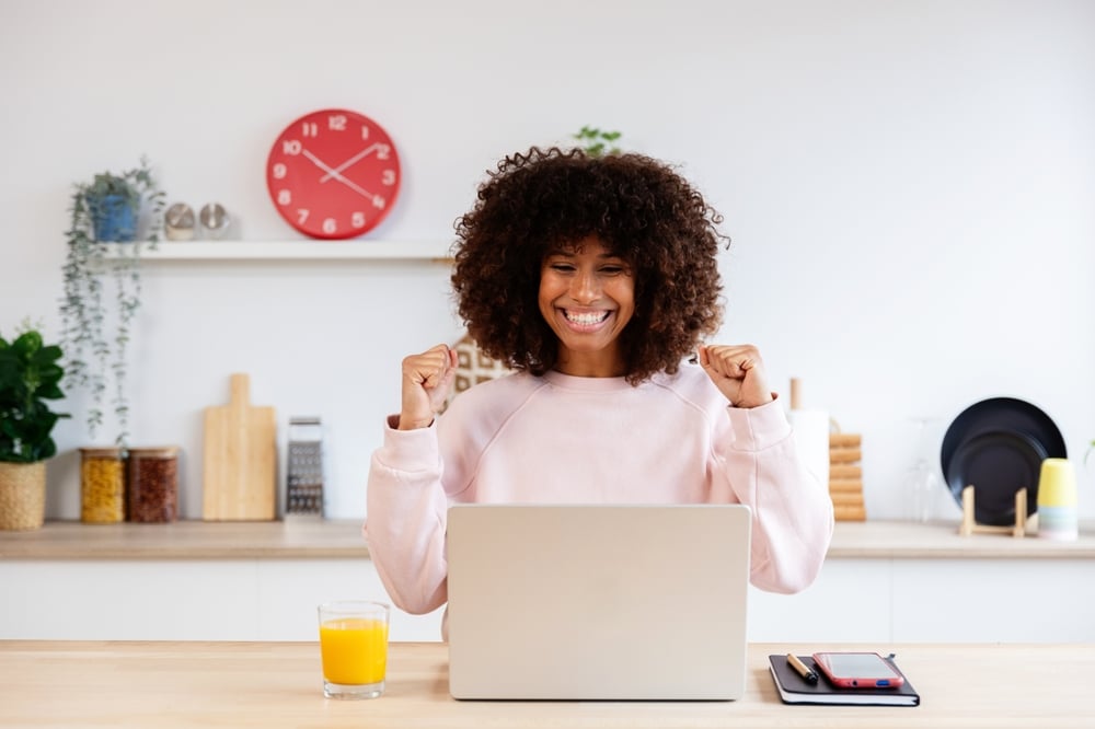 Télétravail réussi : femme joyeuse devant son PC portable. Jeune femme noire aux cheveux bouclés, radieuse, célèbre une réussite professionnelle devant son PC. Jus d'orange et horloge rouge en arrière-plan.