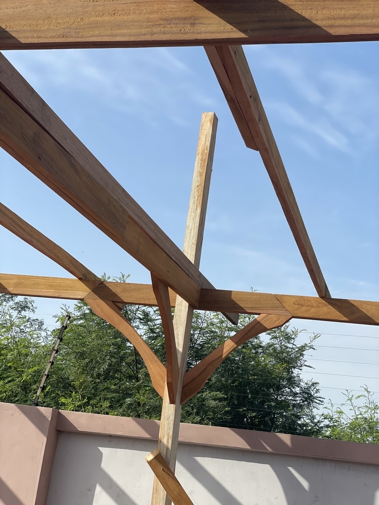 Charpente bois de pergola traditionnelle sous ciel bleu Détail de la charpente en bois massif d'une pergola. Poteau central et poutres soutenus par des courbes élégantes, ciel bleu.